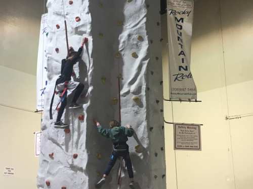 Two climbers ascend a rock wall indoors, with colorful holds and safety harnesses visible.