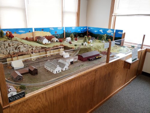 A detailed model train display featuring buildings, trees, and a scenic backdrop of hills and clouds.