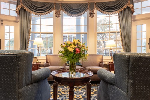 Cozy living room with blue armchairs, a wooden table, and a vase of flowers in front of large windows.