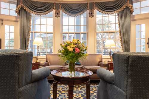 Cozy living room with blue armchairs, a wooden table, and a vase of flowers in front of large windows.