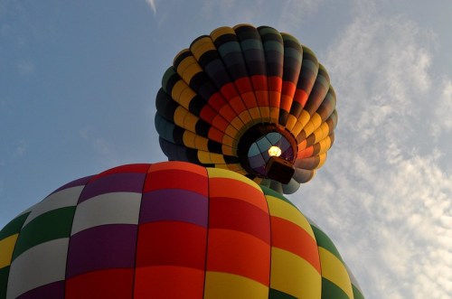Two colorful hot air balloons against a blue sky, one above the other, with vibrant patterns and a soft cloud backdrop.