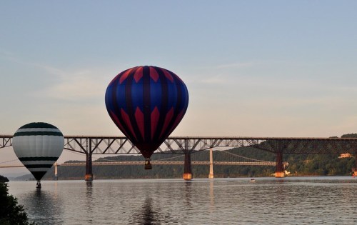 Two hot air balloons float over a calm river, with a bridge visible in the background under a clear sky.