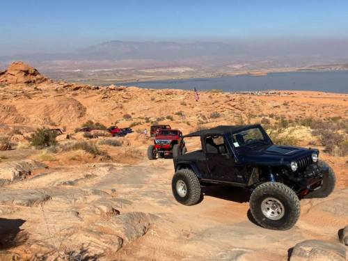 Three off-road vehicles navigate a rocky terrain overlooking a lake and distant mountains under a clear sky.