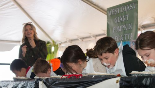 Children compete in a pumpkin pie-eating contest under a tent, with a woman smiling in the background.