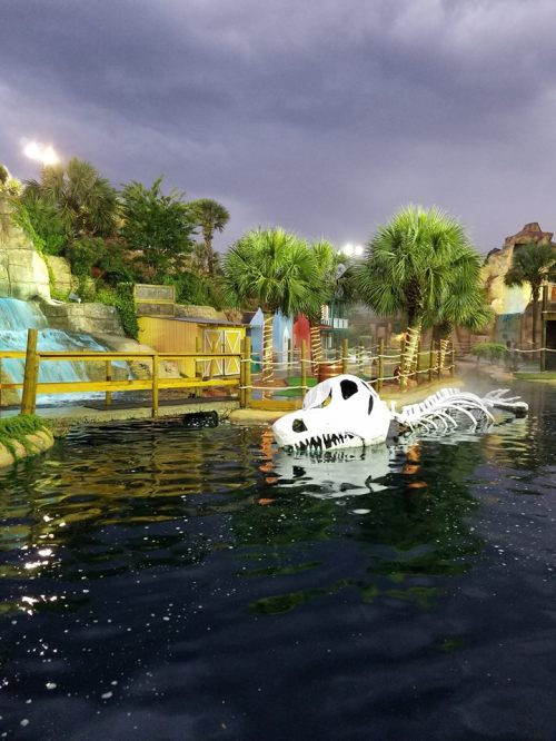 A large dinosaur skeleton floats in a pool surrounded by palm trees and a cloudy sky.