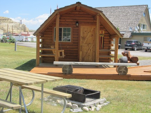 A small wooden cabin with a porch, surrounded by grass, a picnic table, and a fire pit in a scenic outdoor setting.