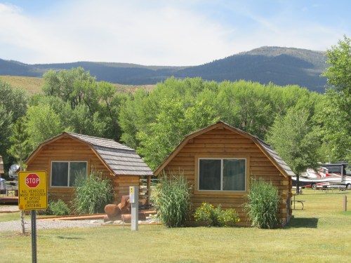 Two wooden cabins surrounded by greenery, with mountains in the background and a stop sign nearby.