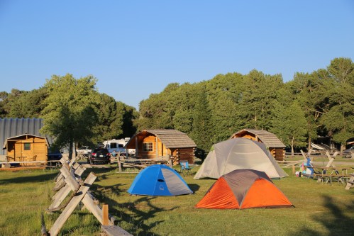 A campsite with colorful tents, wooden cabins, and trees under a clear blue sky.