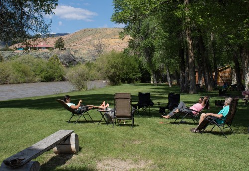 People relaxing in lawn chairs by a river, surrounded by trees and hills under a clear blue sky.