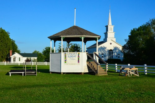 A gazebo with a concert sign, surrounded by green grass, with a church and trees in the background under a clear blue sky.