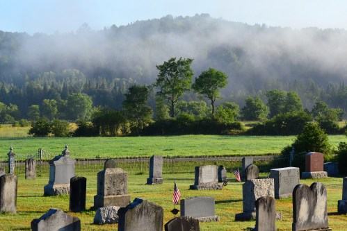 A serene cemetery with gravestones, surrounded by lush greenery and misty hills in the background.
