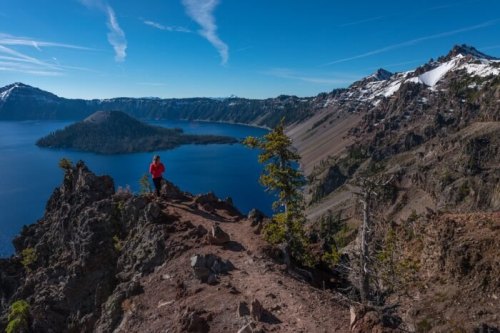 A hiker stands on a rocky ledge overlooking a blue lake surrounded by mountains and trees under a clear sky.