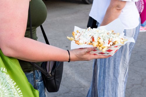 A person holds a tray of loaded fries topped with cheese and sauce, while another person stands nearby.