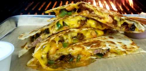 Three cheesy quesadillas filled with beef, peppers, and tomatoes, served with a side of sauce, against a grill backdrop.