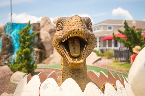 A dinosaur sculpture emerging from an egg at a mini-golf course, with a blue waterfall and trees in the background.