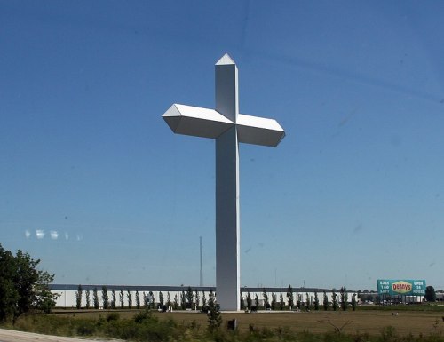 A large white cross stands tall against a clear blue sky, surrounded by open land and a distant billboard.