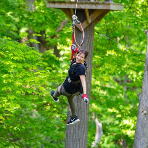 A person joyfully zip-lining through a lush green forest, wearing safety gear and smiling at the camera.