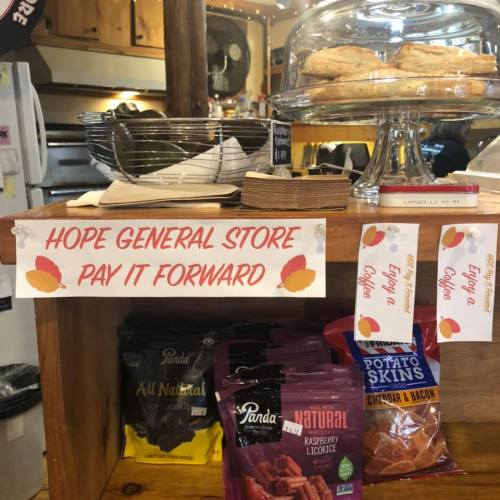 A cozy general store display with snacks, a sign reading "Hope General Store Pay It Forward," and a pastry under a glass dome.