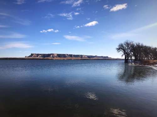 A serene lake scene with a clear blue sky, distant cliffs, and bare trees along the water's edge.