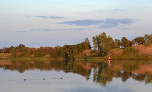 Serene lake at sunset, reflecting trees and hills under a soft, colorful sky.