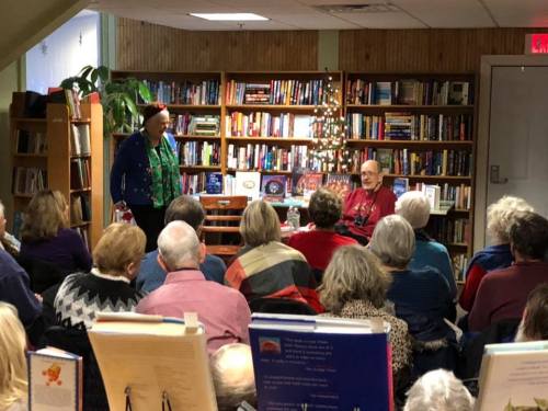 A speaker stands in front of an audience in a cozy library, with bookshelves filled with books in the background.