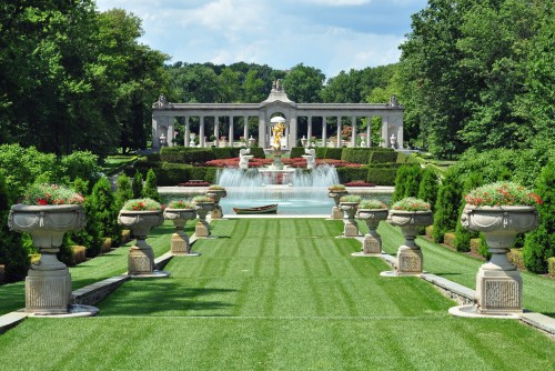A landscaped garden with a fountain, manicured lawns, and decorative urns, framed by trees and a grand structure in the background.