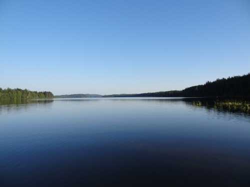 A calm lake reflecting a clear blue sky, surrounded by lush green trees and distant hills.