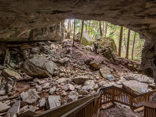 View from inside a cave, showing rocky terrain and trees outside, with wooden stairs leading down into the cave.