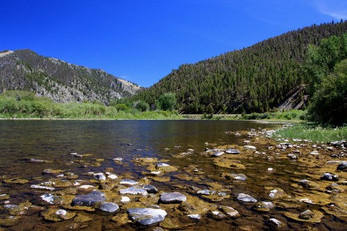 A serene river scene with clear water, rocky shore, and lush green mountains under a bright blue sky.