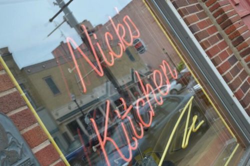 Neon sign reading "Nick's Kitchen" in a window, with a brick wall and reflections of buildings in the background.