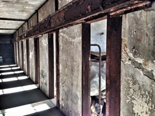 Interior of an old, abandoned building with rusty beams, cracked walls, and a bed in a dimly lit space.
