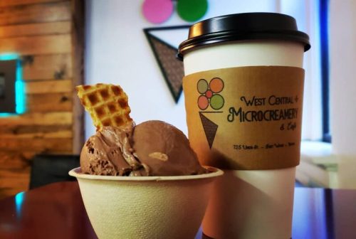 A cup of chocolate ice cream with a waffle cone piece beside a coffee cup, set on a wooden table.