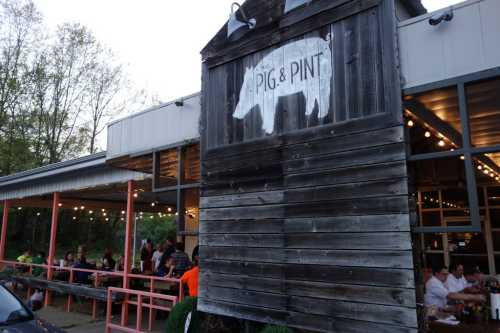 Exterior of a rustic restaurant called "Pig & Pint," with outdoor seating and string lights in the evening.