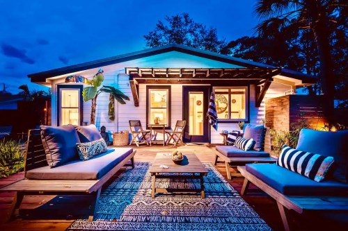 A cozy house at dusk, featuring a deck with seating, plants, and warm lighting, creating a welcoming atmosphere.