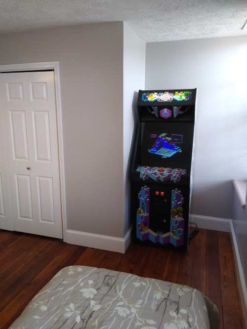 A retro arcade game machine stands in a corner of a room with light gray walls and wooden flooring.