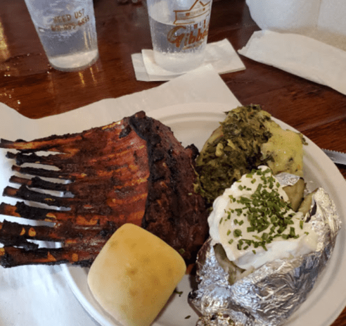 A plate with ribs, a baked potato topped with sour cream and chives, collard greens, and a roll, served on a wooden table.