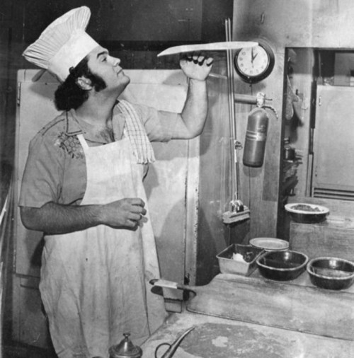 A chef in a kitchen inspects a large knife while standing near a scale and bowls of ingredients.