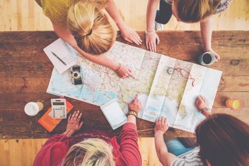 Group of people gathered around a table, studying a large map with notebooks and drinks nearby.