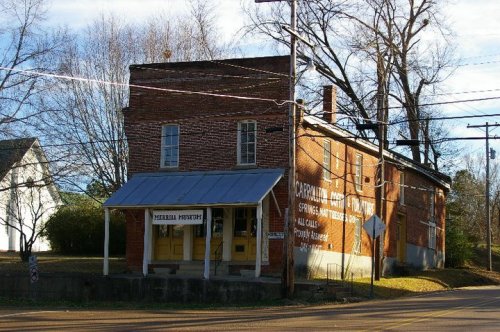 Historic brick building with a porch, featuring a sign for "Merrill Museum" and surrounded by trees and power lines.