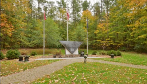 A memorial site surrounded by trees, featuring flags and a triangular monument on a stone pathway.