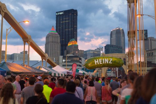 Crowd walking on a bridge with a large pickle sign and city skyline in the background during an event at dusk.