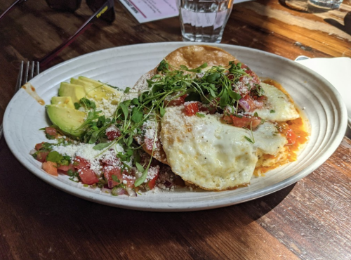 A plate of huevos rancheros with eggs, salsa, avocado slices, and fresh herbs on a wooden table.
