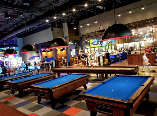 A vibrant entertainment venue featuring pool tables, arcade games, and a lively dining area in the background.