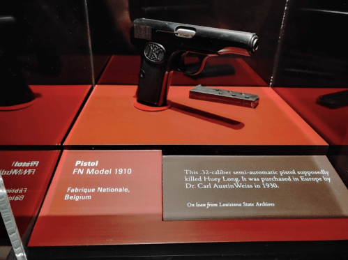 A FN Model 1910 pistol displayed in a museum case, with a plaque detailing its history and origin.