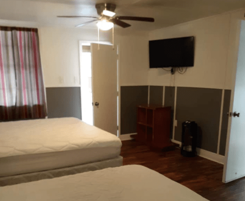 A simple hotel room with two beds, a TV on the wall, a fan, and a door leading outside.