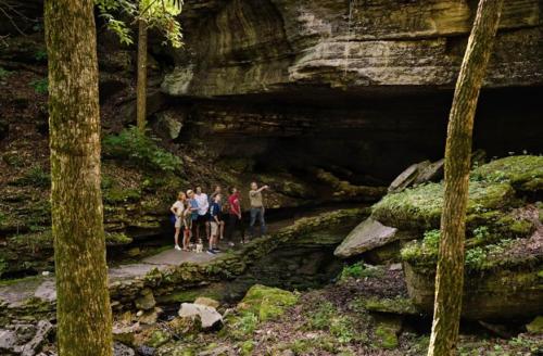 A group of five people stands near a rocky cave entrance in a lush, green forest.