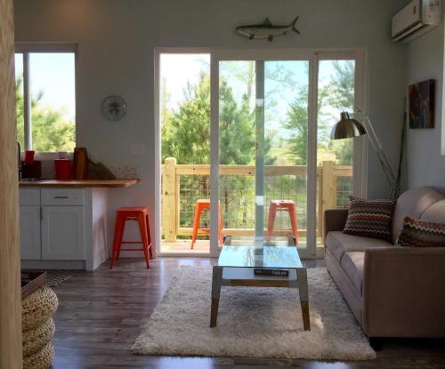 Cozy living space with a sofa, coffee table, and kitchen area, featuring large windows and a view of greenery outside.