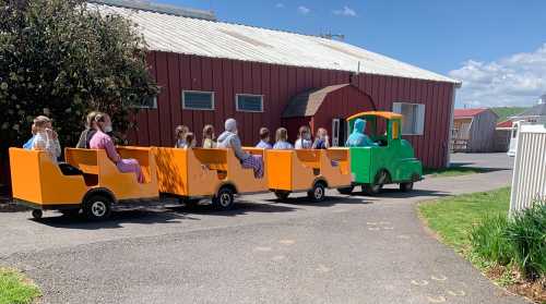 A colorful miniature train with children riding in orange and green cars on a sunny day near a red building.