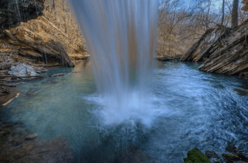 A serene waterfall cascading into a clear blue pool, surrounded by rocky terrain and bare trees.