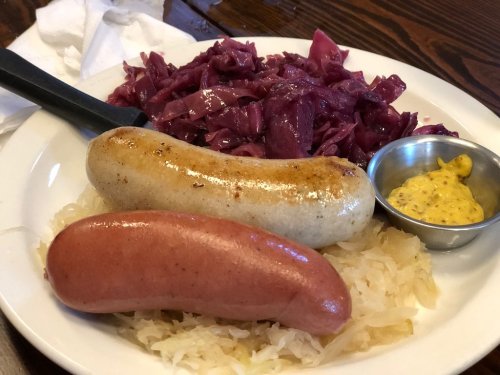 A plate with two sausages, one white and one pink, served with sauerkraut and red cabbage, alongside a small bowl of mustard.
